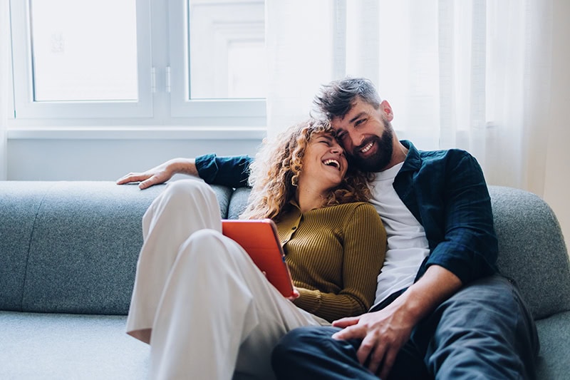 A happy couple cuddling closely on a grey sofa near a window with sheer curtains.