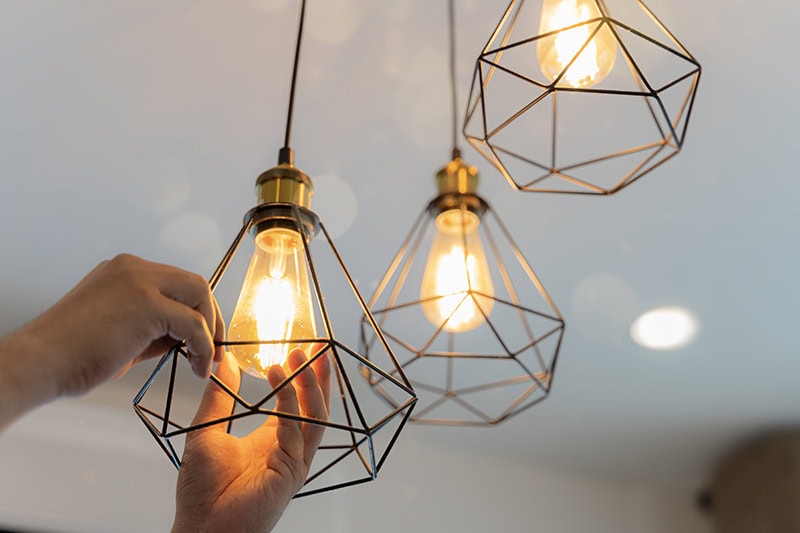 A close-up, low-angle view of someone's hands adjusting or installing a lit Edison-style light bulb inside a modern, black geometric wire cage pendant fixture.