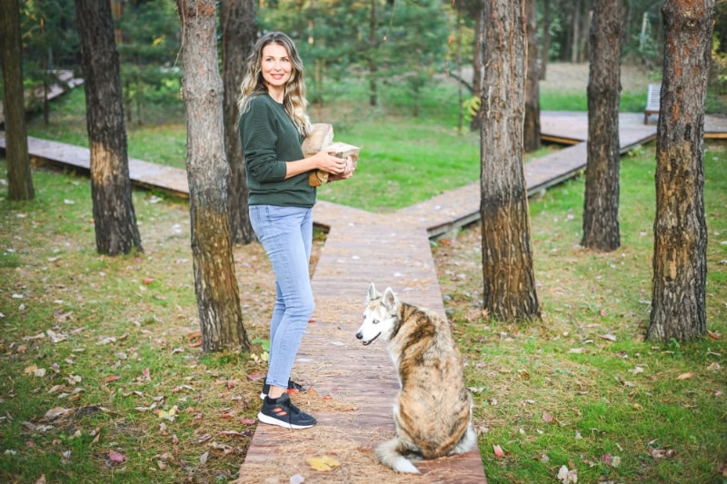 Woman holding her dog and walking with another on a wooden path in the woods.